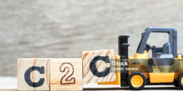 A miniature yellow forklift carrying a wooden block with the letter 'C', next to two other blocks forming the text 'C2C', on a wooden surface with a blurred background.