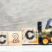 A miniature yellow forklift carrying a wooden block with the letter 'C', next to two other blocks forming the text 'C2C', on a wooden surface with a blurred background.
